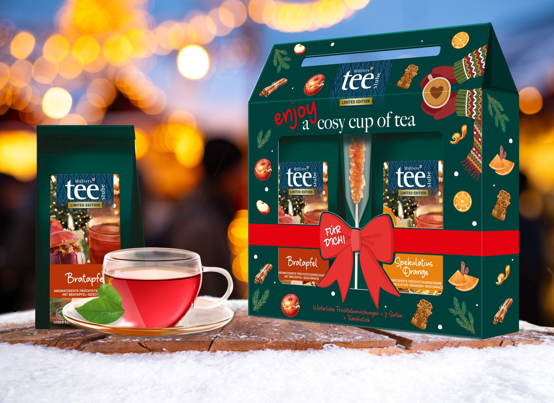 Ein festliches Tee-Geschenkset und eine Tasse roter Tee stehen auf einem Tisch mit einem verschwommenen, warm beleuchteten Hintergrund. Die Verpackung ist mit weihnachtlichen Motiven und Etiketten versehen.